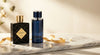 Two luxury perfume bottles on a marble surface surrounded by white petals and a golden ribbon, evoking a high-end fragrance editorial.